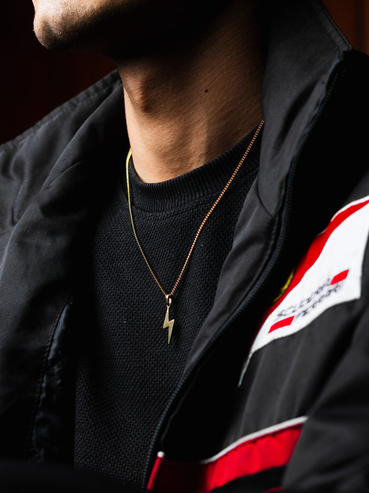 Model styling a gold Lightning pendant necklace on top a black jacket with red and white accents.